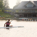 young man paddling away from us in kayak with yacht club building in background