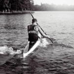 grayscale image of young man paddling away from us in kayak