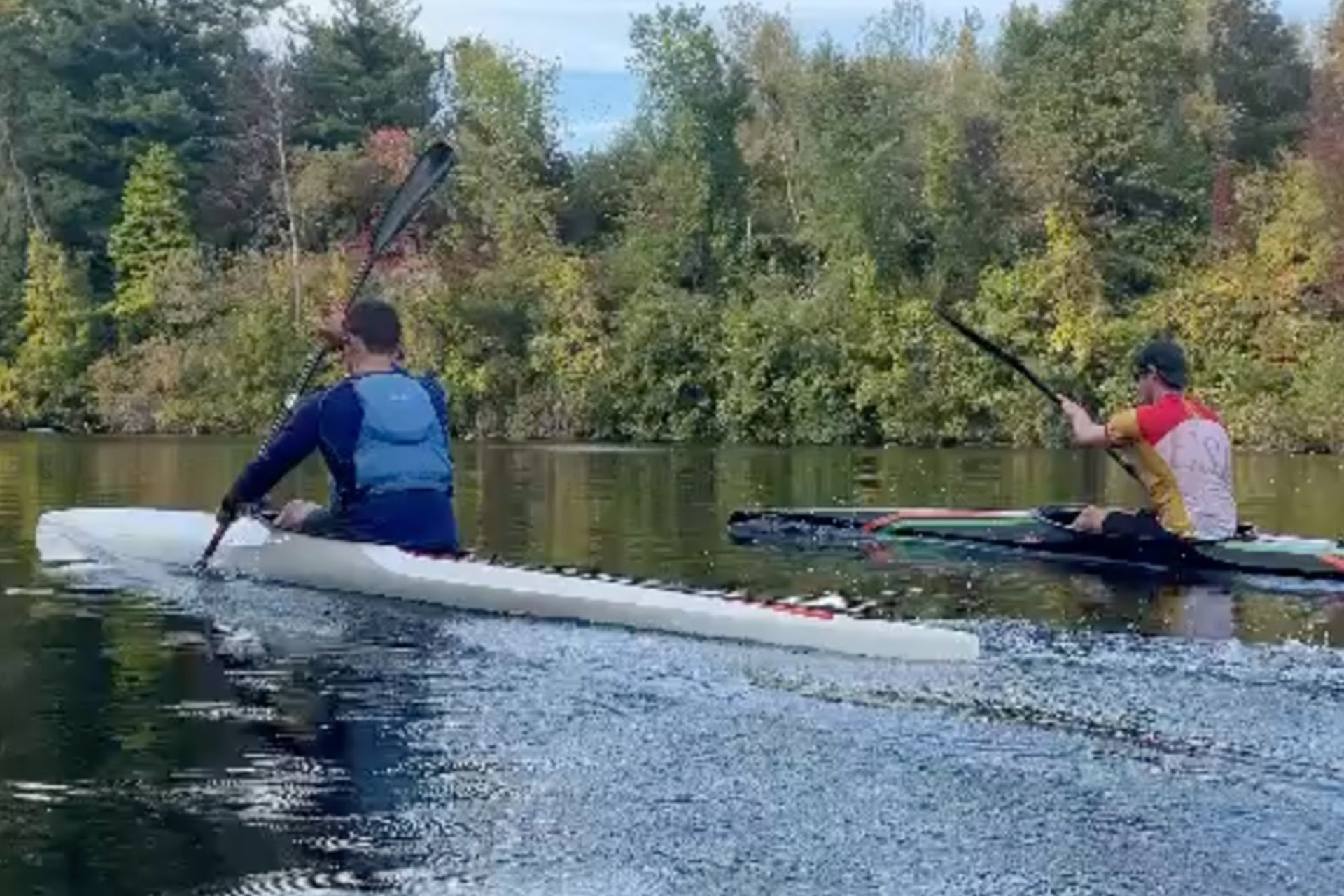 two young men paddling kayaks on a lake with autumn trees in background